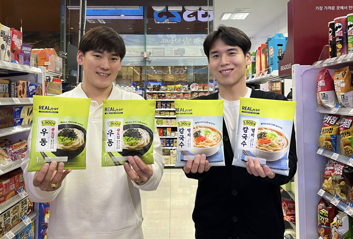 편의점에서 두 남성이 REALprice 브랜드의 우동과 칼국수 라면 제품을 들고 미소 짓고 있는 모습