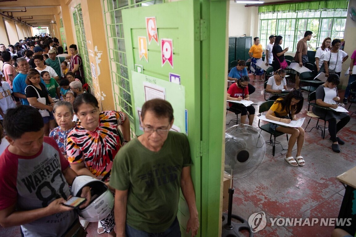 사람들이 복도에 길게 줄을 서 있고, 교실 안에서는 사람들이 책상에 앉아 서류를 작성하고 있는 모습