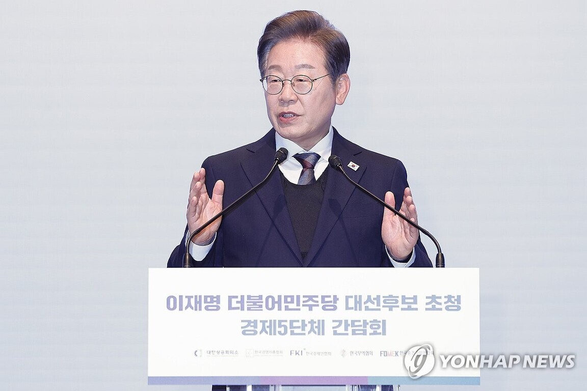 정장을 입은 남성이 연단에서 두 손을 들어 연설하고 있다. 연단에는 '이재명 더불어민주당 대선후보 초청 경제5단체 간담회'라는 문구가 적혀 있다.