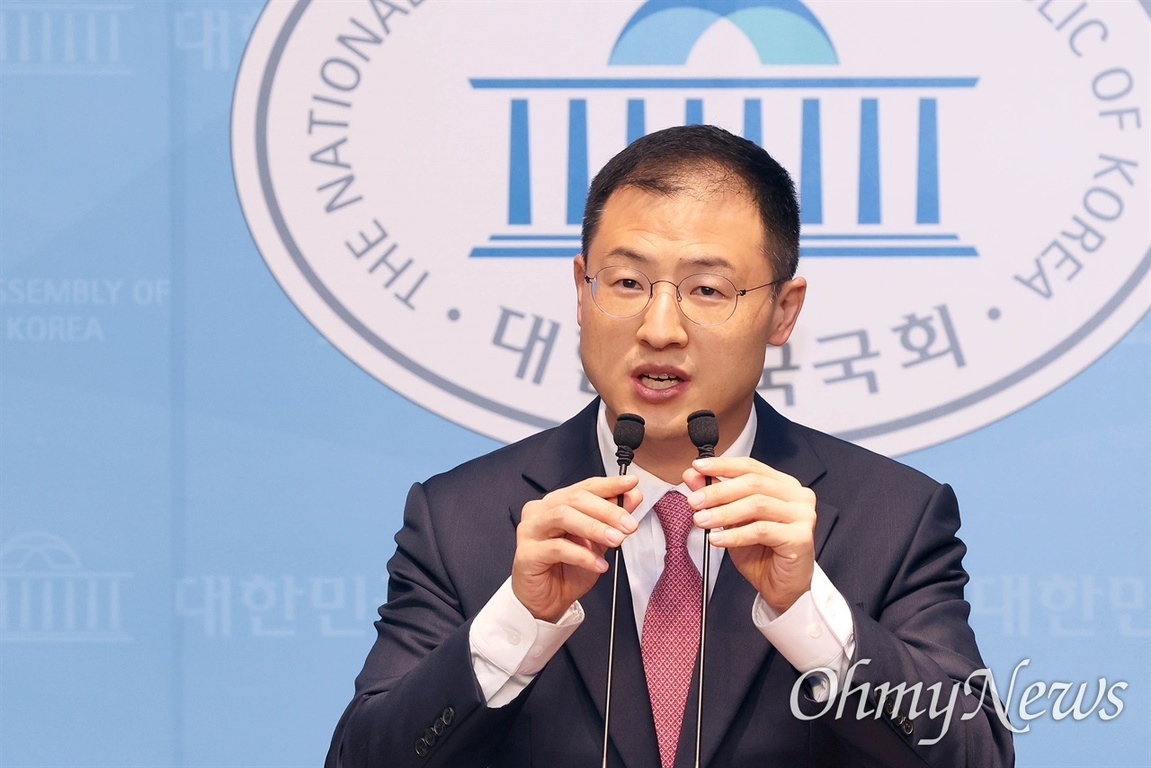 정장과 넥타이를 착용한 남성이 국회 로고 앞에서 마이크를 잡고 발언하고 있다.