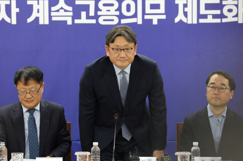 정장 차림의 남성 세 명이 책상에 앉아 있고, 가운데 남성이 일어나 인사하는 모습이다. 배경에는 한글로 된 파란색 현수막이 걸려 있다.