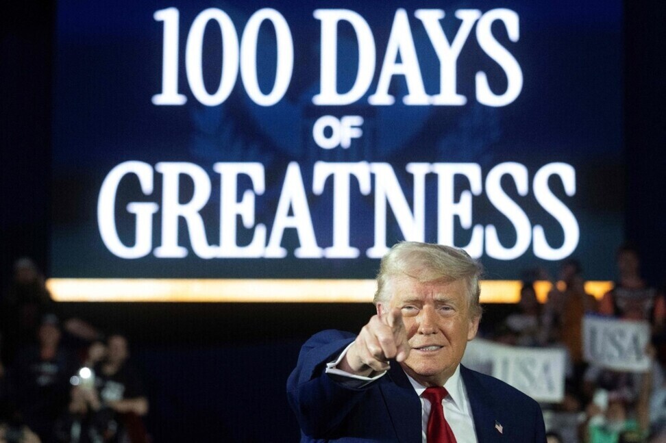 한 남성이 무대 앞에서 손가락으로 가리키고 있으며, 배경에는 '100 DAYS OF GREATNESS'라는 대형 문구가 보인다.