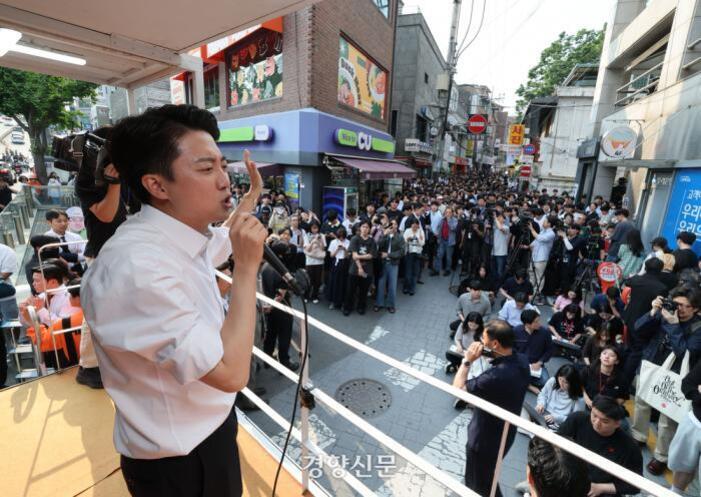 한 남성이 마이크를 들고 연단에서 연설을 하고 있으며, 많은 사람들이 길거리에서 이를 지켜보고 있다. 배경에는 편의점과 상점들이 보인다.