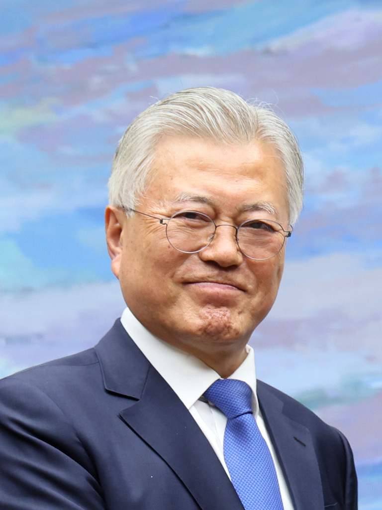 안경을 쓴 노년의 남성이 정장을 입고 미소를 짓고 있다. 배경은 파스텔톤의 추상화이다.