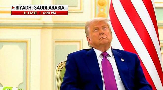 한 남성이 정장과 보라색 넥타이를 착용하고 의자에 앉아 있으며, 배경에는 미국 국기와 'RIYADH, SAUDI ARABIA'라는 뉴스 자막이 보인다.