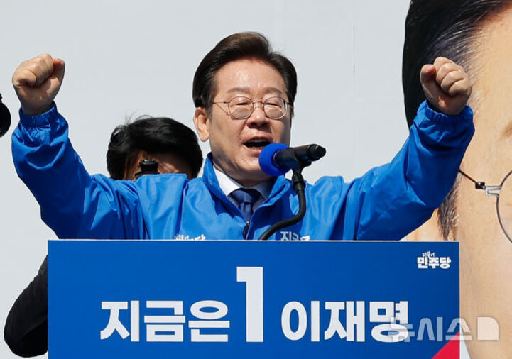 파란색 점퍼를 입은 남성이 연단에서 두 팔을 들어 올리며 연설하고 있다. 연단에는 '지금은 1 이재명'이라는 문구가 적혀 있다.