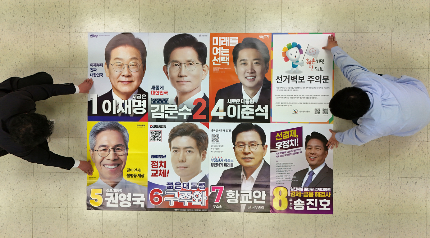 두 명의 남성이 바닥에 여덟 명의 대통령 후보 포스터와 선거 벽보 주의문을 정렬하고 있는 모습. 각 포스터에는 후보자의 사진, 이름, 정당 및 번호가 표시되어 있다.