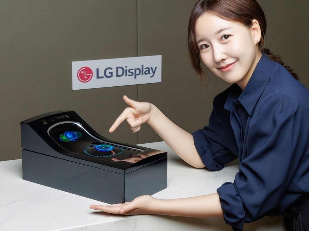 여성이 LG Display 로고가 있는 배경 앞에서 곡면 디스플레이 장치를 가리키며 미소 짓고 있다.