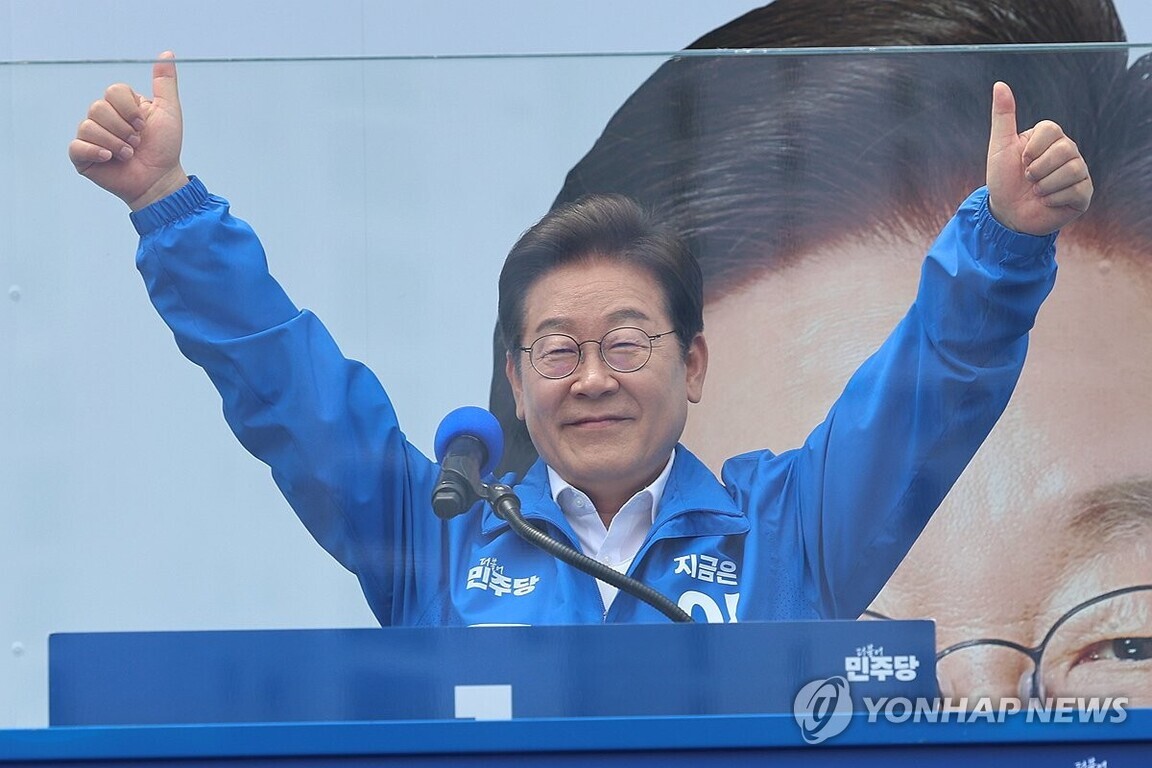 파란색 점퍼를 입은 남성이 연단에서 두 손을 들어 엄지손가락을 치켜세우고 있다. 마이크와 파란색 배경, 그리고 배경에는 확대된 얼굴 사진이 보인다.