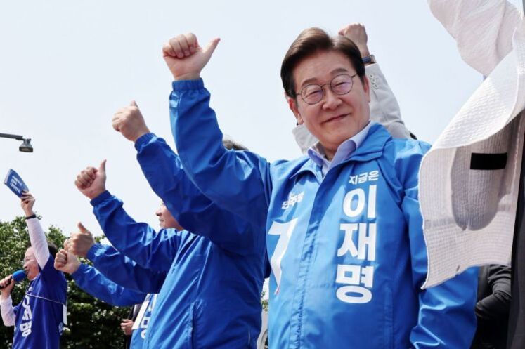 여러 명의 사람들이 파란색 점퍼를 입고 엄지손가락을 들어 올리며 단체로 포즈를 취하고 있다. 앞쪽 인물의 점퍼에는 '이재명'이라는 글자가 적혀 있다.