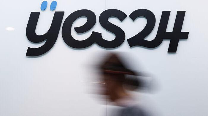 흰 벽에 'yes24'라는 검은색 로고가 크게 보이고, 앞을 흐릿하게 지나가는 사람의 실루엣이 있다.
