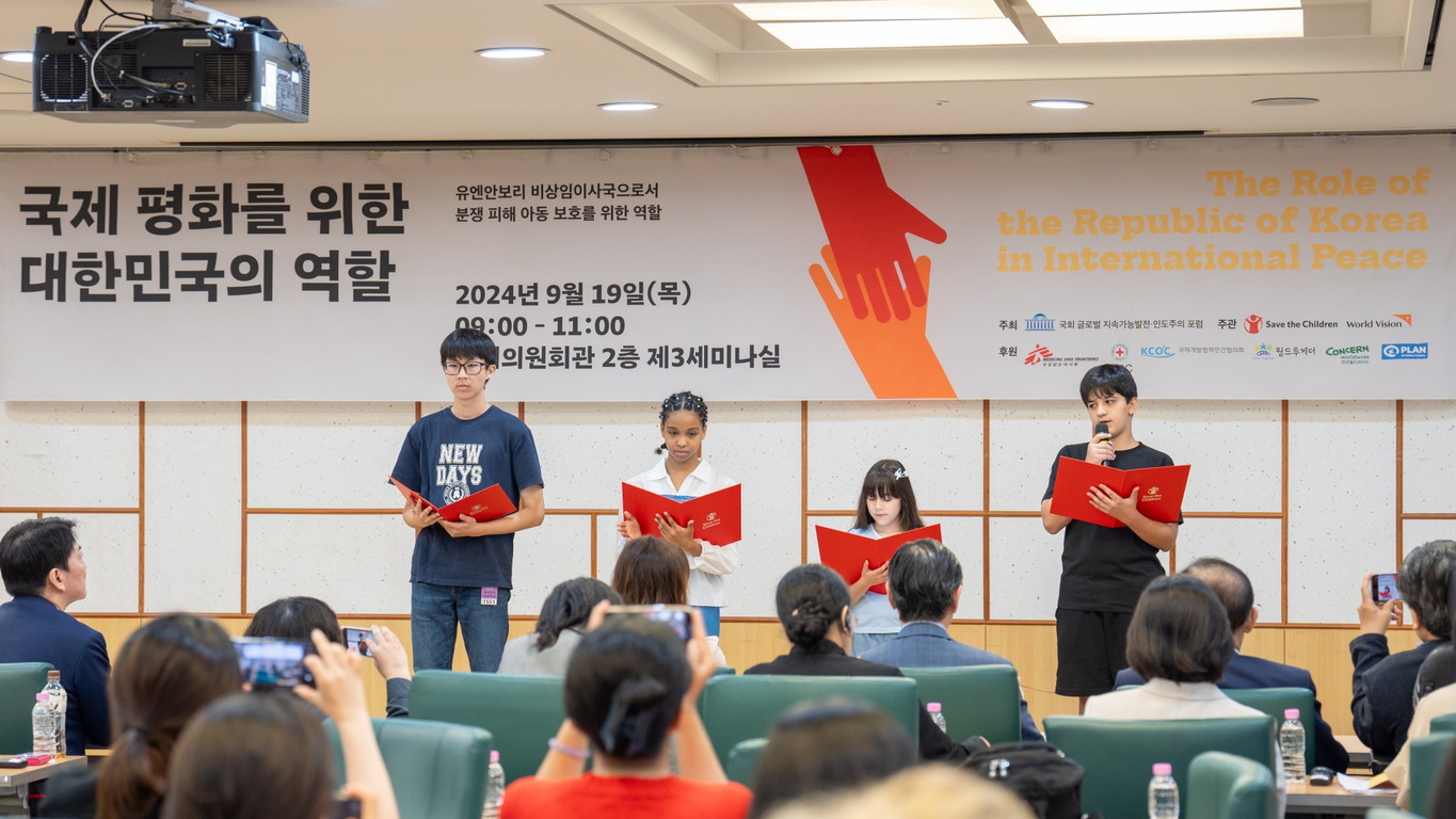 국제 평화를 위한 대한민국의 역할 세미나에서 네 명의 청소년이 단상에 서서 발표를 하고 있으며, 청중들이 이를 지켜보고 있다. 배경에는 행사명과 일시, 주최 및 후원 기관이 적힌 현수막이 걸려 있다.