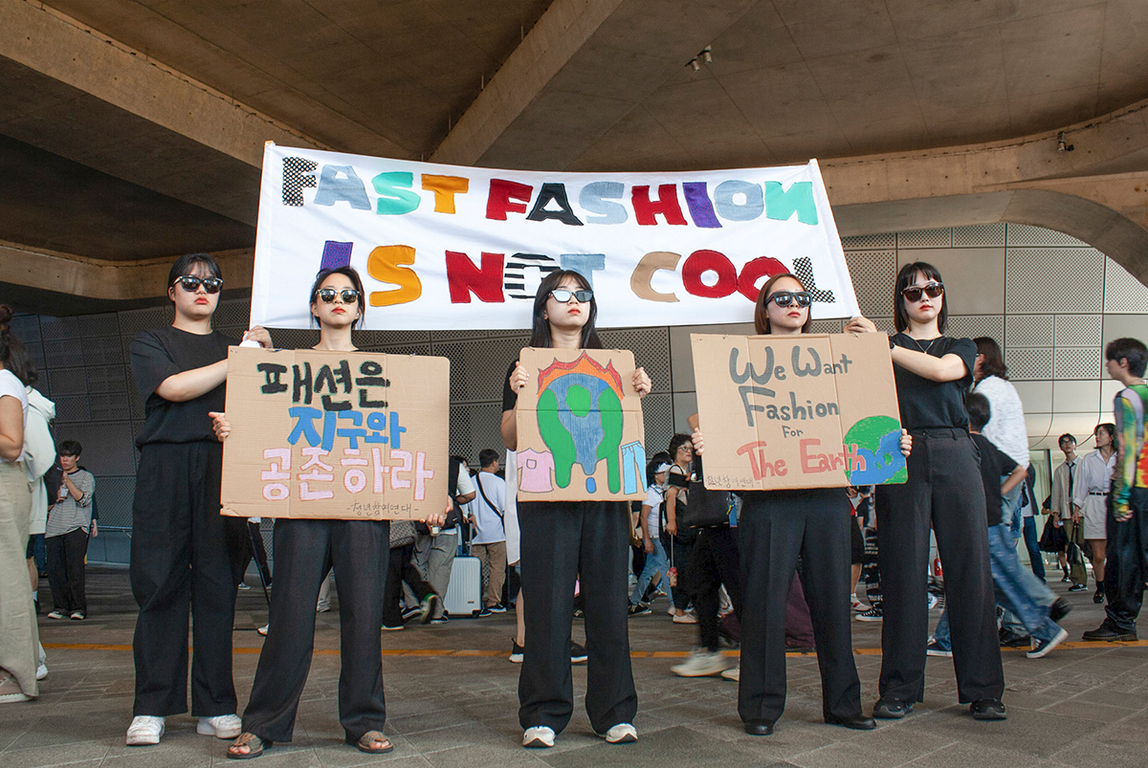네 명의 젊은 여성이 'FAST FASHION IS NOT COOL'이라고 적힌 현수막과 환경 보호 메시지가 담긴 피켓을 들고 패스트패션 반대 시위를 하고 있다.