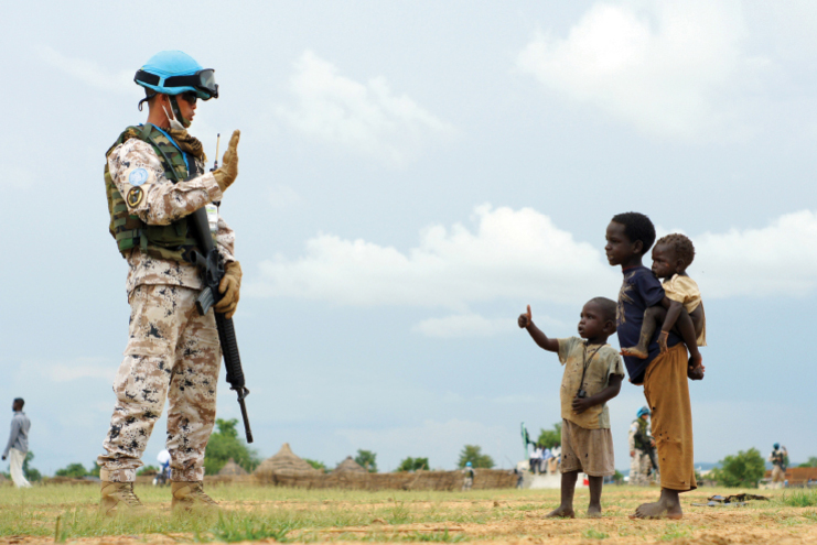 파란 헬멧을 쓴 군인이 세 명의 어린이와 마주 서서 손을 들어 인사하고 있다. 어린이 중 한 명은 엄지손가락을 들어 보이고, 다른 두 명은 그 옆에 서 있다. 배경에는 초원과 하늘이 보인다.