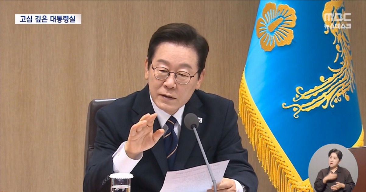 정장 차림의 남성이 마이크 앞에서 종이를 들고 회의 중 발언하고 있다. 뒤에는 파란색 바탕에 금색 무늬가 있는 깃발이 보이고, 화면 오른쪽 아래에는 수어 통역사가 있다.