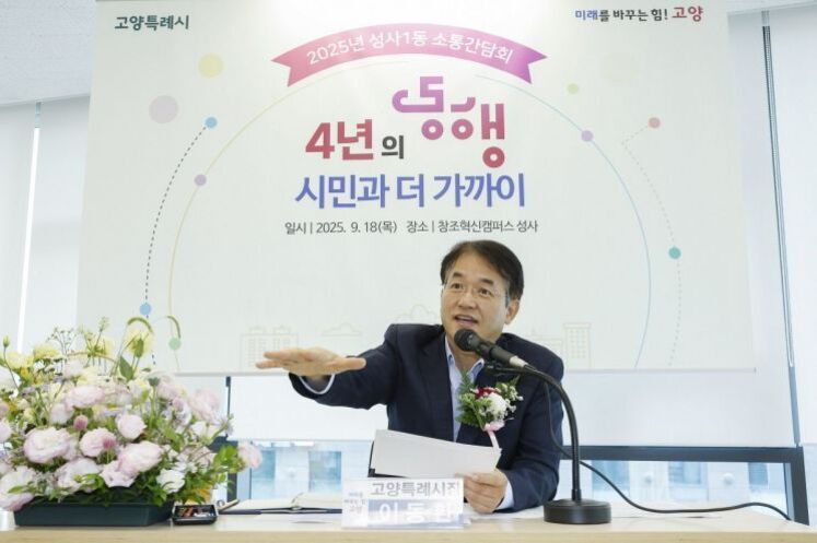 정장 차림의 남성이 마이크 앞에서 손을 내밀며 발언하고 있다. 배경에는 '4년의 동행 시민과 더 가까이'라는 문구와 행사 정보가 적힌 현수막이 걸려 있고, 테이블 위에는 꽃다발과 명패가 놓여 있다.