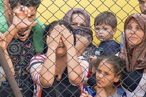 철망 울타리 뒤에 여러 명의 남녀와 아이들이 모여 있으며, 한 여성은 두 손으로 얼굴을 가리고 있다. 사람들은 걱정스럽고 불안한 표정을 짓고 있다.