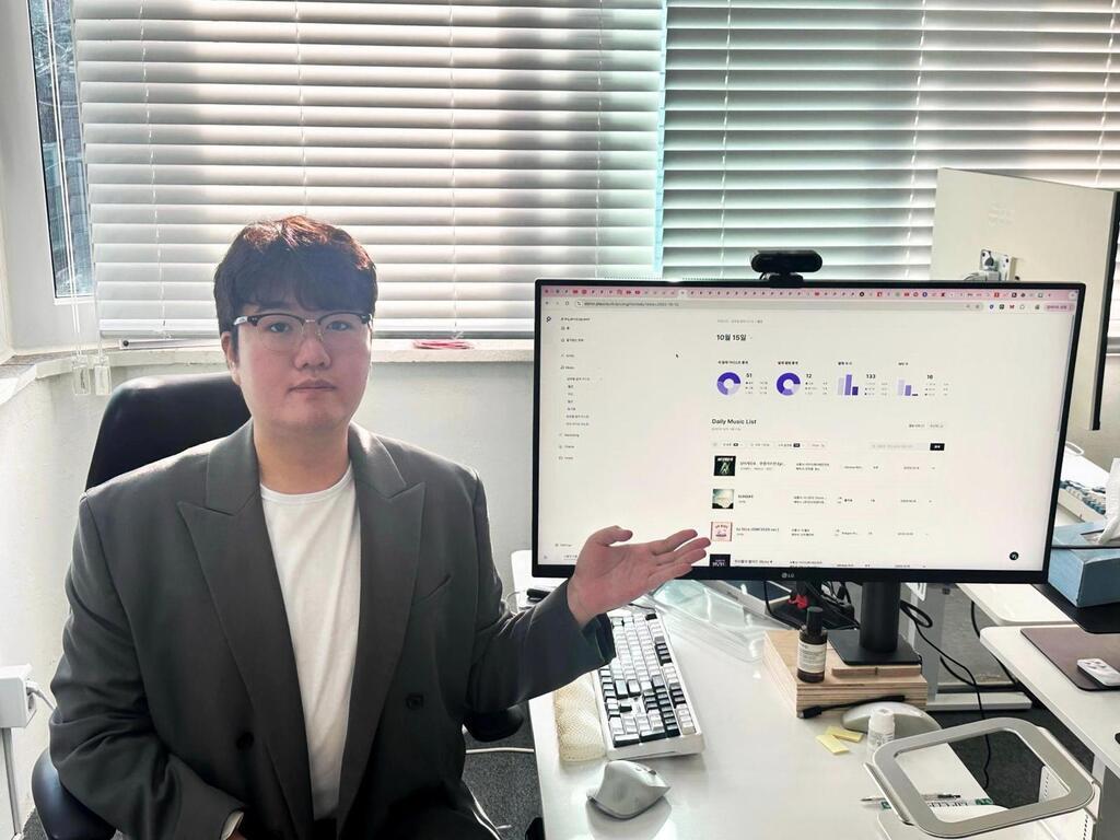 안경을 쓴 남성이 사무실 책상에 앉아 모니터 화면을 손으로 가리키고 있다. 모니터에는 그래프와 리스트가 표시되어 있다.