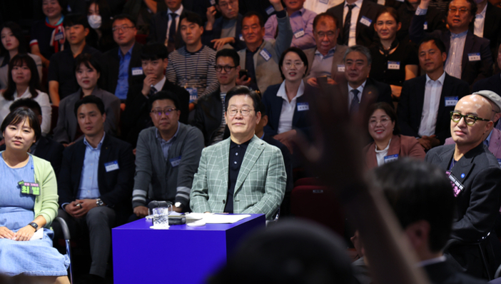 많은 사람들이 앉아 있는 토론회 또는 공개 행사 현장으로, 가운데에 앉은 남성이 주목받고 있다. 앞쪽에는 파란색 테이블이 놓여 있다.