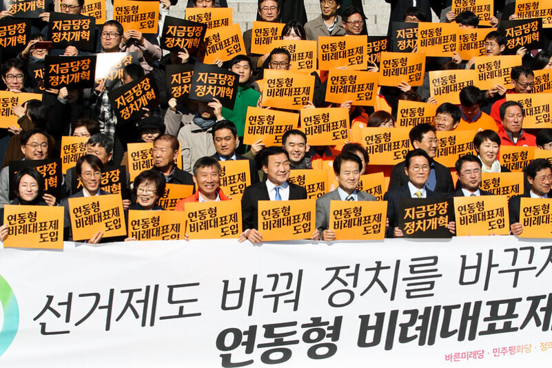 여러 명의 사람들이 '연동형 비례대표제 도입', '지금당장 정치개혁'이라고 적힌 주황색 팻말을 들고 선거제도 개혁을 촉구하는 집회에 참여하고 있다. 앞에는 '선거제도 바꿔 정치를 바꾸자 연동형 비례대표제 도입'이라는 대형 현수막이 펼쳐져 있다.