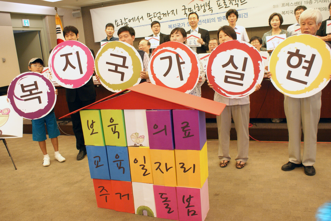 여러 명의 사람들이 '복지국가 실현'이라는 글자가 적힌 원형 팻말을 들고 있고, 앞에는 '보육', '의료', '교육', '일자리', '주거', '돌봄' 등 복지 관련 단어가 적힌 집 모양의 구조물이 놓여 있다. 배경에는 현수막이 걸려 있다.