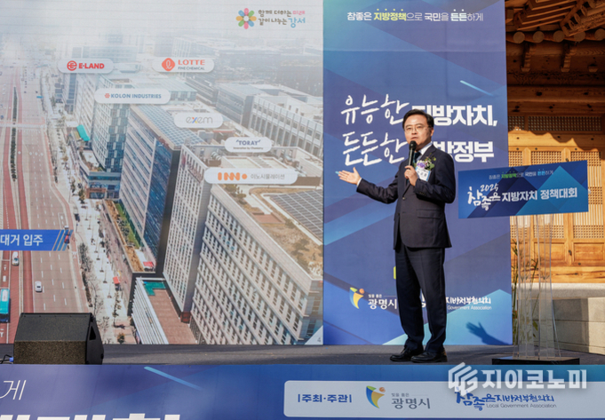 정장을 입은 남성이 무대에서 마이크를 들고 발표하고 있다. 배경에는 대형 건물들이 보이는 도시 사진과 '유능한 지방자치, 든든한 지방정부'라는 문구가 적힌 대형 패널이 있다.