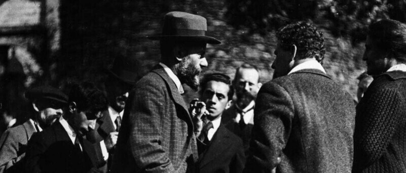 여러 명의 남성이 야외에서 모여 대화를 나누고 있는 흑백 사진. 한 남성은 중절모를 쓰고 있다.