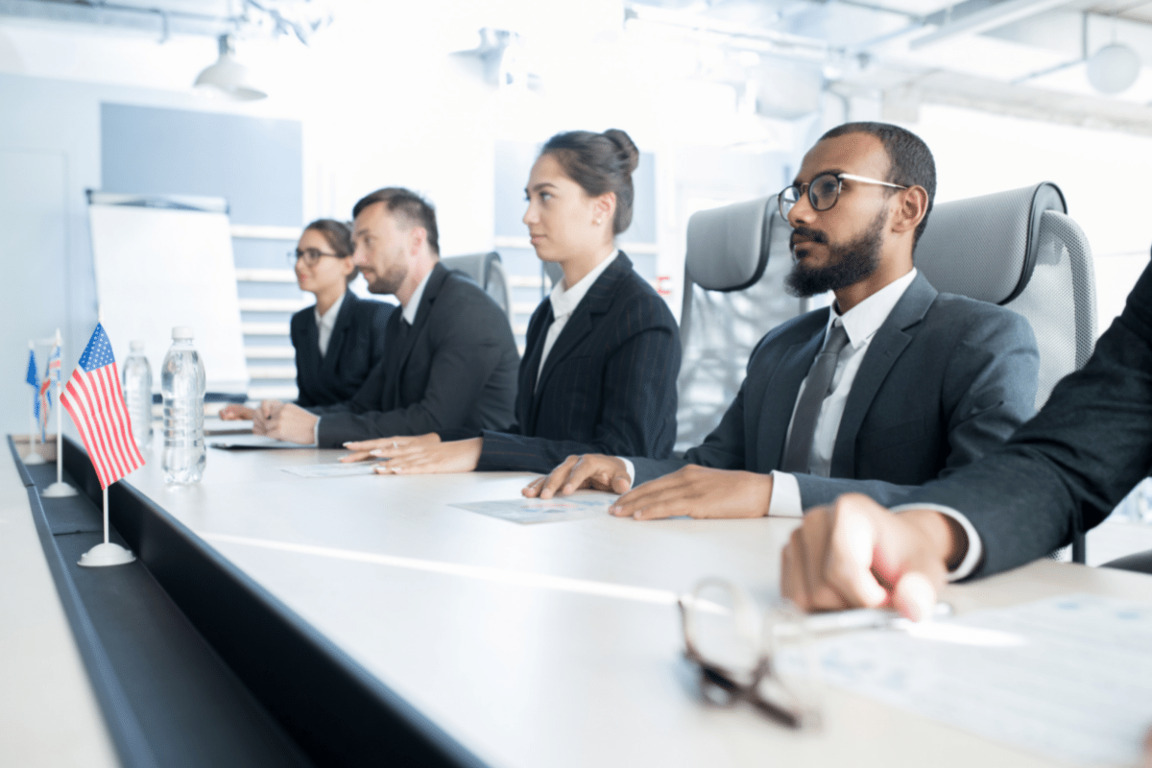 국제 경제 협력의 현재와 미래: 글로벌 전략과 사례 분석 3 정장 차림의 남녀 네 명이 회의실 책상에 앉아 있으며, 책상 위에는 미국 국기와 물병이 놓여 있다.